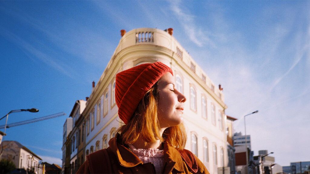 A woman stands in the sunlight with her eyes closed outside a building